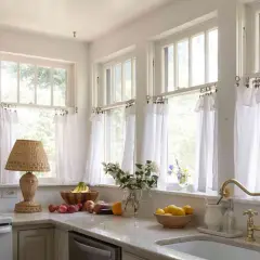 A bright kitchen with white diy cafe curtains framing the windows. A wicker lamp, fruit bowls, and a brass faucet add charm.