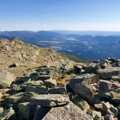 Vista panorámica de los picos de la Sierra de Guadarrama, con rocas cubiertas de líquenes en primer plano y un valle con un lago al fondo.