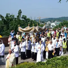 Procesja z monstrancją w słoneczny dzień, kiedy jest Boże Ciało. Ludzie w strojach liturgicznych i świeckich idą ulicą.