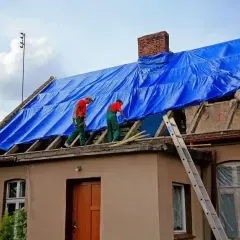 Pracownicy w zielonych ogrodniczkach i czerwonych koszulkach zabezpieczają dach niebieską plandeką podczas przebudowy domu.