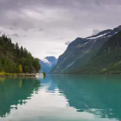 Turkusowa woda odbija skaliste góry i lasy. Statek cumuje przy brzegu, w oddali widać lodowiec. Piękne fiordy w Norwegii.