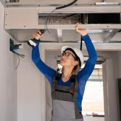 Une femme en casque et lunettes, avec une perceuse, installe un faux plafond pour un système de climatisation split gainable.