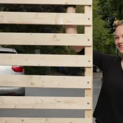 Junge Frau zeigt Daumen hoch neben selbstgebautem Lattenrost. Ein Auto ist im Hintergrund zu sehen.