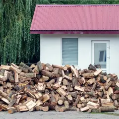 Duża sterta drewna opałowego przed białym budynkiem, z zielonym lasem w tle. Odległość budynku od lasu jest niewielka.