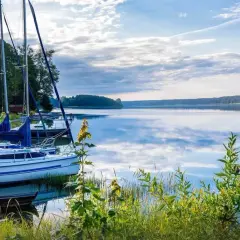 Żaglówki przy pomoście na spokojnym jeziorze. W tle las, a na wodzie odbijają się chmury. Mazury ciekawostki: idealne miejsce na relaks i żeglowanie.