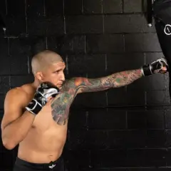 A tattooed man in boxing gloves performs a punching bag workout, his fist connecting with the bag.