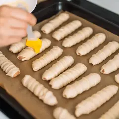 Ręka smaruje pędzelkiem parówki zawinięte w ciasto, przygotowując je do pieczenia. To prosty przepis na parowance.
