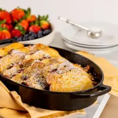 En ugnsform med gyllene fattiga riddare, pudrad med florsocker. En skål med färska jordgubbar och blåbär står i bakgrunden.