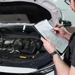 Mechanik sprawdza listę kontrolną przy otwartej masce samochodu. Dowiedz się, ile kosztuje badanie techniczne samochodu.