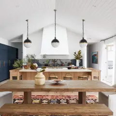 A modern lake house interior featuring a rustic wooden dining table, plush white chairs, and a sleek navy kitchen island.
