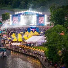 Tłumy ludzi na festiwalu Rap Stacja, scena z ekranami i dymem, żółte parasole, łódź na wodzie.