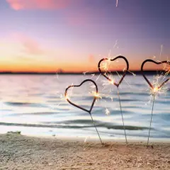 Drei Herz-Wunderkerzen funkeln am Strand. Ein romantischer Silvester am Meer mit einem farbenprächtigen Himmel.