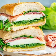 Dos bocadillos italianos apilados, rellenos de mozzarella fresca, prosciutto, rúcula y pesto, sobre pan rústico.