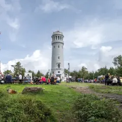 Ludzie odpoczywają przy wieży widokowej i maszcie telekomunikacyjnym. W oddali widać wielką sowę na tle chmur.