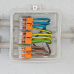 Bezpieczne przyłącze elektryczne z przewodami w kolorach niebieskim, żółto-zielonym i brązowym, połączonymi złączkami WAGO 221 w białej puszce.