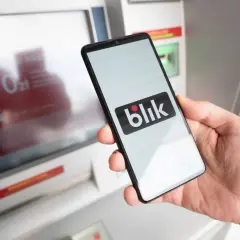 Dłoń trzyma smartfon z logo Blik przed bankomatem. Blik działa w Polsce i kilku innych krajach Europy.