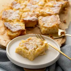 Ein saftiger Apfelkuchen vom Blech, bestreut mit Puderzucker und Mandelblättchen. Ein Stück liegt auf einem Teller, bereit zum Genuss.