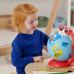 Un bambino di 5 anni con capelli rossi esplora il mondo con un globo interattivo, circondato da matite colorate.