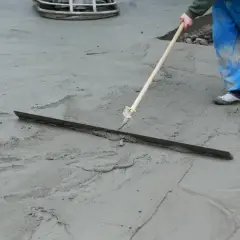 Pracownik w niebieskich ogrodniczkach wygładza świeży beton długą pacą. Trwa wygrzewanie posadzki.