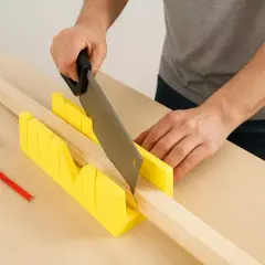 Un bricoleur utilise une boîte à onglets jaune pour une coupe plinthe précise. Un mètre ruban et un crayon sont à proximité.