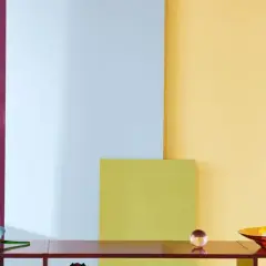 Stolik z lampą i misą owoców na tle kolorowych paneli, jakby pomalowanych farbą malarską rozpuszczalną w wodzie.