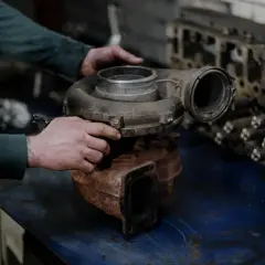 Mechanik trzyma turbosprężarkę, przygotowując się do jej wymiany krok po kroku. Obok leży głowica silnika.