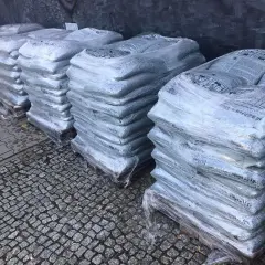 Stosy worków z asfaltem na paletach, owinięte folią, na kamiennym podłożu.