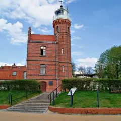 Ceglana latarnia morska w Ustce z czerwoną dachówką i zielonymi żywopłotami.