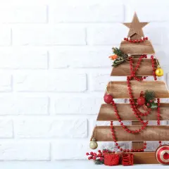 Drewniana choinka z desek DIY, ozdobiona czerwonymi koralami, bombkami i prezentami, stoi na tle białej ceglanej ściany.