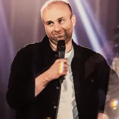 Marcin Zbigniew Wojciech w swoim stand-upie, trzyma mikrofon, a światła sceniczne tworzą tęczowe refleksy.
