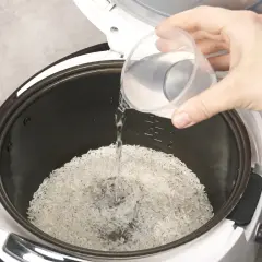 Beim Reis kochen wie viel Wasser? Eine Hand gießt Wasser in den Reiskocher über den Reis.