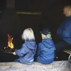 Två barn och en vuxen sitter vid en eld. En mysig paus under vandring med barn.
