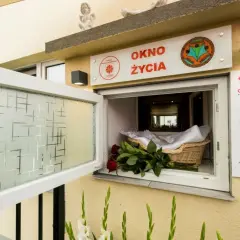 Okno życia, bezpieczne miejsce dla noworodków, jest otwarte. W koszyku czeka otulone kocykiem niemowlę, a obok bukiet czerwonych róż.