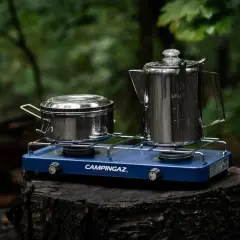 Niebieska kuchenka turystyczna Campingaz z dwoma palnikami, na której stoją metalowy garnek i kawiarka. Idealna na biwak.