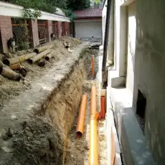 Trwa budowa drenażu wokół domu. W wykopie leżą nowe pomarańczowe rury i stare, zniszczone rury.