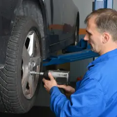 Mechanik w niebieskim kombinezonie dokręca śruby kół aluminiowych przy podniesionym samochodzie.