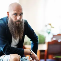 Łysy z brodą w eleganckiej marynarce i podartych dżinsach, z niebieskimi oczami, spogląda wprost na widza.