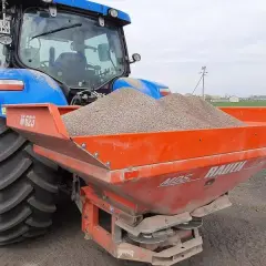 Niebieski traktor z przyczepionym rozsiewaczem napełnionym wapnem, gotowy do pracy na polu.