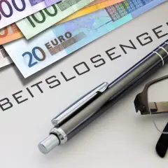 Euro-Scheine liegen neben einem Stift und einer Brille auf einem Dokument mit der Aufschrift "ABFINDUNG ARBEITSLOSENGELD".