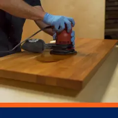 Main tenant des mains gantées, une ponceuse électrique effectue le lustrage bois d'un plateau.