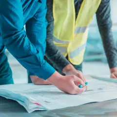 Deux hommes examinent des plans. L'un, en chemise bleue, pointe un détail avec un stylo. L'autre, en gilet jaune, pose ses mains sur les documents. Un constat huissier avant travaux est en cours.
