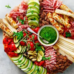 Tabla de antipasti italiani con prosciutto, salami, mozzarella, aguacate, tomates, pimientos y calabacín a la parrilla, acompañada de pesto y pan.