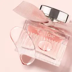 Szklana buteleczka L'eau de Chloe z różową wstążką i kroplami perfum na delikatnym, różowym tle.