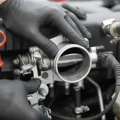 Mechanik w czarnych rękawiczkach sprawdza przepustnicę. Objawy jej awarii mogą być różne, np. nierówna praca silnika.