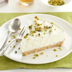 Rebanada de tarta de coco mousse, cubierta con coco rallado y pistachos picados, sobre plato blanco con cubiertos.
