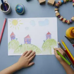 Dziecko rysuje kredkami domki na wzgórzach, pokazując, jak się rysuje dom. Słońce i chmury dopełniają krajobraz.