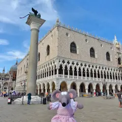 Une souris en robe rose devant l'entrée du Palais des Doges à Venise, avec la colonne de Saint-Marc et la basilique Saint-Marc en arrière-plan.
