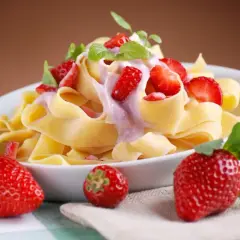 Pappardelle z kremowym sosem i kawałkami truskawek, ozdobione listkami mięty. Obok talerza leżą świeże truskawki.