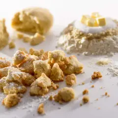 Knusprige Streusel mit gemahlenen Mandeln, bestäubt mit Puderzucker. Im Hintergrund Teig und Zutaten für eine köstliche Leckerei.