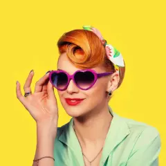 Une femme au style rockabilly, cheveux coiffés en chignon, porte des lunettes violettes en forme de cœur et un foulard coloré.